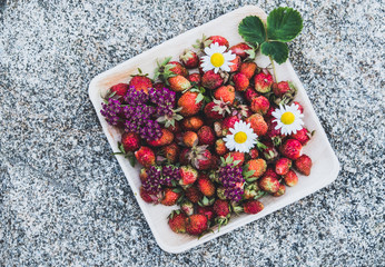 Fresh red strawberries in a wooden plate. Strawberry is a source of vitamin C. This berry is a natural aphrodisiac. Collage with wildflowers on the background of natural granite stone.