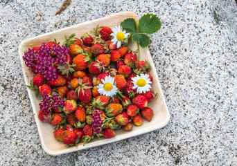 Fresh red strawberries in a wooden plate. Strawberry is a source of vitamin C. This berry is a natural aphrodisiac. Collage with wildflowers on the background of natural granite stone.