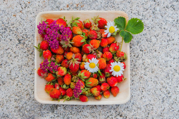 Fresh red strawberries in a wooden plate. Strawberry is a source of vitamin C. This berry is a natural aphrodisiac. Collage with wildflowers on the background of natural granite stone.