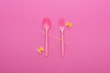 pink plastic spoon and fork, spaghetti and two uncooked farfalle pasta isolated on pink
