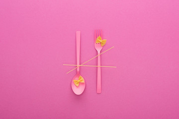 pink plastic spoon upside down and fork, spaghetti and two uncooked macaroni isolated on pink