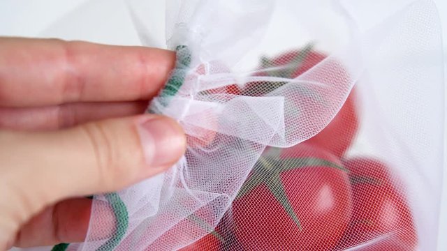 female hand touches ripe branch of cherry tomato lies in a reusable reuse bag on an isolated white background. to deal with disposable plastic bags close up mesh texture