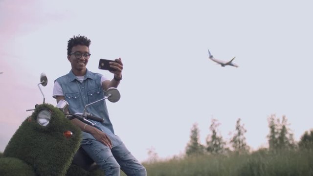 Photo of a handsome excited young business man walking outdoors on scooter take a selfie by camera. Plane on the background