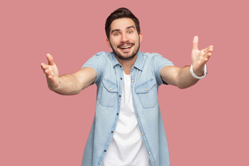 Fototapeta premium Portrait of happy handsome bearded young man in blue casual style shirt standing with raised arms, want to hug and looking at camera with toothy smile. indoor studio shot, isolated on pink background.