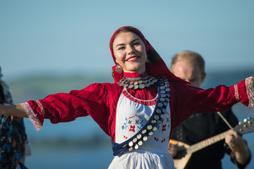 Young smiling woman in traditional russian clothes dancing on the field - a man playing balalaika...