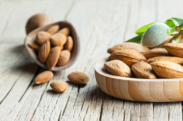 Almond nuts in wooden shovel, almonds with shell in bamboo bowl on wooden background with green fresh raw almonds on almond tree branch.