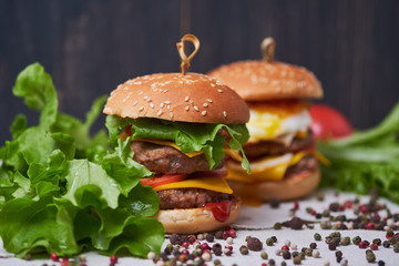 Two big classic american cheeseburger with two chops. Cheeseburgers on a linen tablecloth, decorated with different types of pepper and lettuce. Dark wooden background.
