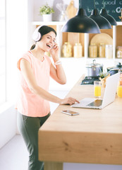 Portrait of a cheerful young woman listening to music with headphones and using laptop computer while standing at the kitchen