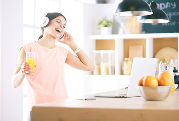 Portrait of a cheerful young woman listening to music with headphones and using laptop computer while standing at the kitchen
