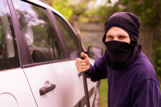 masked thief standing near the car and trying to smash the window with cowbar