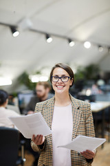 Waist up portrait of smiling businesswoman posing in office holding documents, copy space