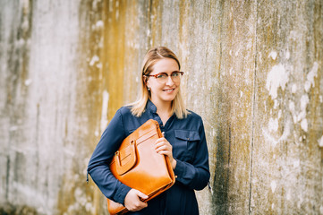 Outdoor portrait of young business woman holding brown leather briefcase, wearing glasses and blue dress