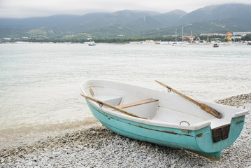 side view of small wooden fishing azure boat on pebble coast black sea beach in bad weather on sea, mountain and resort town background, horizontal stock photo image