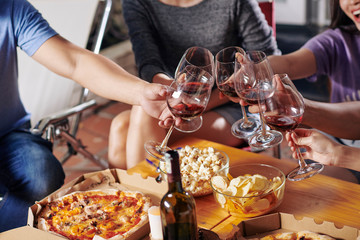 Group of friends clinking wine glasses over table with pizza and snacks