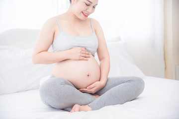 Pregnant woman sitting on bed and holding hands on her belly with smiles. Pregnancy, maternity, preparation and expectation concept.