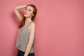 Fototapeta premium Redhead girl stands slightly sideways to the camera, touches the back of her head and smiles at the camera