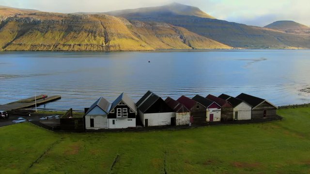 Aerial Antique Shacks in a Row at Coast of Lake, Drone Turn to View Mountains, Streymoy Island, Faroe Islands