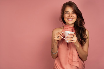 Young dark-haired woman with pink cup in her hands smiles at the camera, pink background