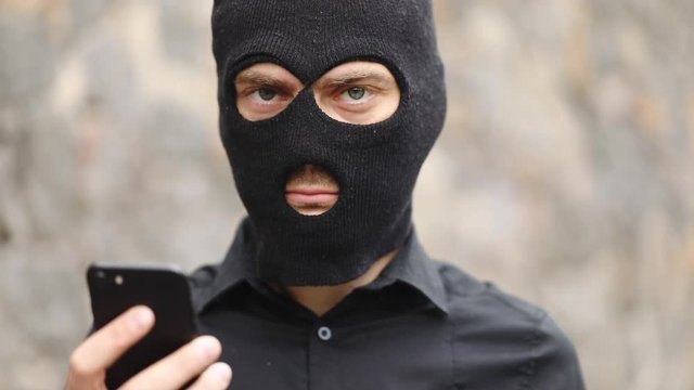 angry criminal in burglar mask looks at modern smartphone and straight at alleged victim slow motion close view