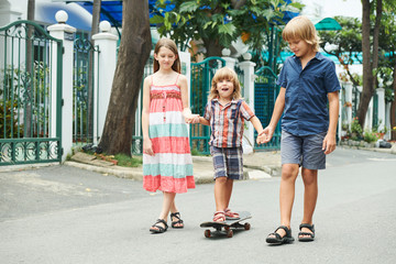 Cheerful kids holding hands of little boy on skateboard riding in the street