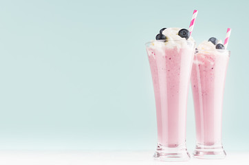 Blueberry milkshake with berry, whipped cream, striped straw in two high transparent glass on white wood table and mint color wall, copy space.