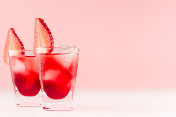 Bright red alcoholic shots in two shot glasses with strawberry slice, ice cubes in trendy modern bar interior in pastel pink color on white wood board.