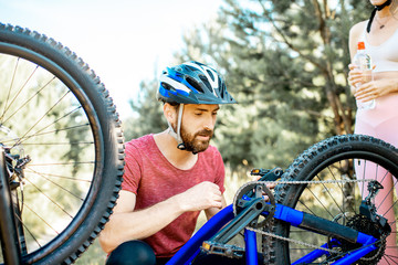 Young man in sportswear repairing bicycle on the forest road during the summer time