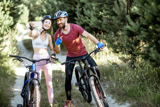 Young sports couple taking selfie photo with smartphone while riding mountain bicycles on the forest road during the summer time