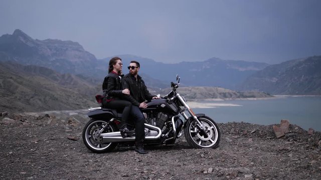 lady and bearded guy in sunglasses sit on modern silver black motorbike against lake surrounded by hills slow motion
