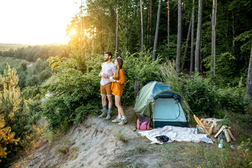 Young and cheerful couple standing together near the tent in the forest, enjoying beautiful...