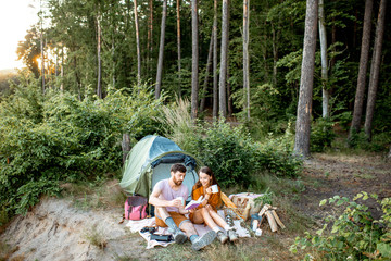 Young and cheerful couple sitting with book near the tent, enjoying the summer vacations in the forest during the sunset