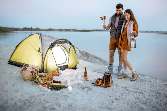 Young couple having fun at the campsite on the beach, standing near the fireplace during the evening