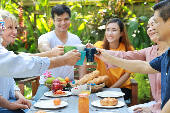 Happy multiethnic family sitting at a breakfast table in backyard outdoor on sunny day with smiling face while everyone clicking glasses.
