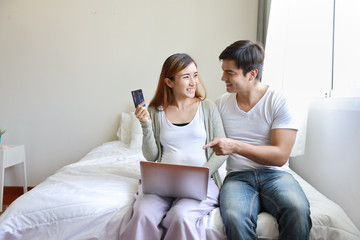 front view young pregnant asian holding credit card for shopping online while using computer with caucasian husband embracing on bed in bedroom with happy smiling face and eyes contract together