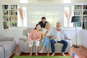full shot of a group of happy elder multiethnic family sitting on grey sofa in living room and watching football or favourite channel with exciting face