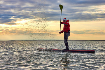 Man paddle (floating) on Stand up paddle board (SUP ) in Santa Claus costume in calm of winter lake...