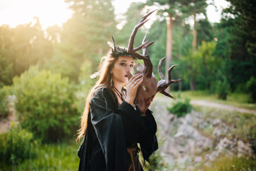 A girl in a terrible mask with horns is standing in nature. A man in a black cloak. The pagan witch conjures. Fantasy image