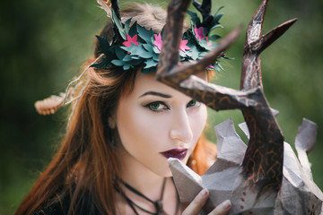 A girl in a terrible mask with horns is standing in nature. A man in a black cloak. The pagan witch conjures. Fantasy image