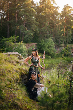 Two beautiful, forest witches stand on a hill with a staff next to the forest. Druids. Fantasy image