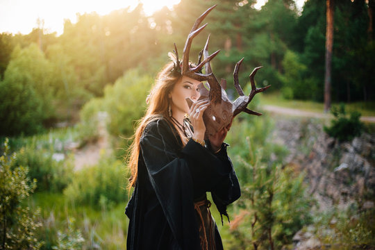 A girl in a terrible mask with horns is standing in nature. A man in a black cloak. The pagan witch conjures. Fantasy image