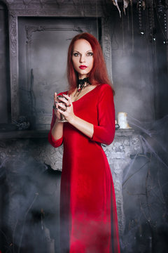 Beautiful red-haired girl with red lips. A model in a red dress with a black necklace in the castle. Vampire style. The image for halloween. Fantasy. Iron cup with wine
