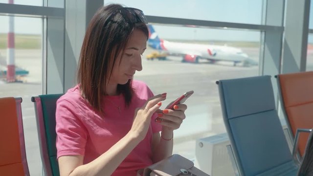 A young beautiful girl sits in the airport waiting room and uses a mobile phone. Woman buying e-ticket, making hotel reservations and checking in online.