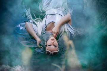 A girl elf in a white dress is floating in the water. Model in a medieval dress with pigtails floating in the lake