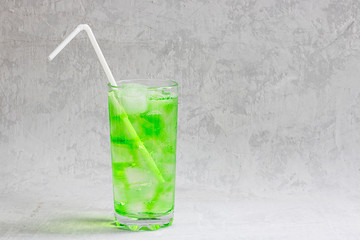 Summer cold green drink with lime, mint and lemon in tall glass with ice and straw and defocused fruit around on grey background. Sparkling beverage, alcohol drink, mojito coctail. Horizontal