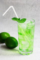Summer cold green drink with lime, mint and lemon in tall glass with ice and straw and defocused fruit around on grey background. Sparkling beverage, alcohol drink, mojito coctail. Vertical