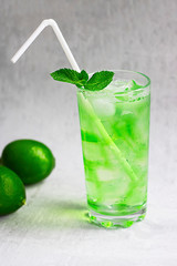 Summer cold green drink with lime, mint and lemon in tall glass with ice and straw and defocused fruit around on grey background. Sparkling beverage, alcohol drink, mojito coctail. Vertical