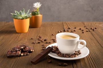 A coffee cup with coffee beans and chocolate on a wooden table with colorful flower pots, selective focus