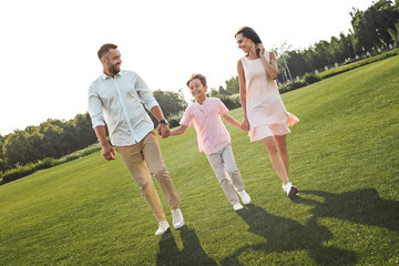 Enjoying morning together. Full length of happy and young family of three holding hands, smiling and walking outdoors in the nature