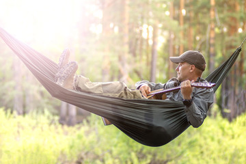 Portrait of young man in hat play guitar and swinging in hammock of weekend morning in camp rest