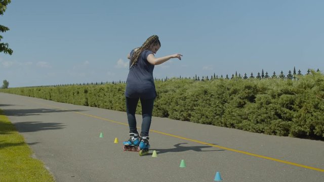 Rear view of active woman inline skating backwards crisscross through cones standing in line on park alley. Skillful female practicing slalom backward bypass cones riding rollerblades in public park.